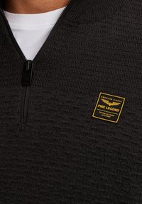 Čierny zipsový fleece pulóver s textúrovaným povrchom, s žltou nášivkou loga PME Legend na ľavej prsnej časti, okrúhlym golierom a zipsovým uzáverom.