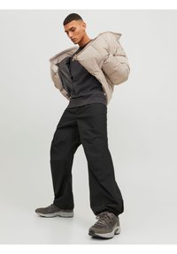 Chaqueta acolchada beige con cremallera, sudadera negra, pantalones negros holgados y zapatillas grises con acentos texturizados y cordones visibles.