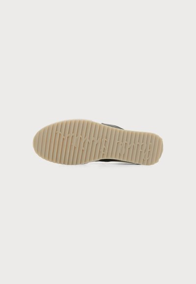 Semelle d'une basket beige avec des rainures horizontales et le nom de la marque en relief visible, présentée sur un fond blanc uni.