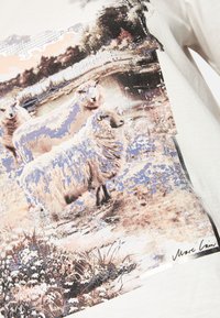 Lichtbeige katoenen T-shirt met een landschapprint van schapen, bomen en een rivier; versierd met zilveren studs bij de halslijn.