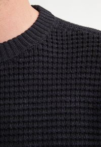 Close-up van een persoon in een zwart wafelgebreid sweater met een geribde ronde halslijn tegen een witte achtergrond.