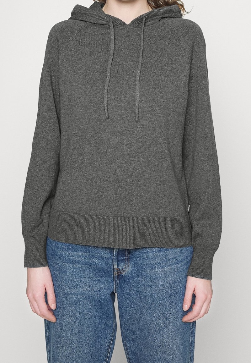 Personne portant un sweat à capuche en maille gris foncé et un jean en denim bleu, debout devant un fond clair uni.