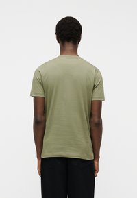 Personne aux cheveux courts et bouclés portant un t-shirt à manches courtes vert olive et un pantalon noir, vue de dos sur un fond uni.