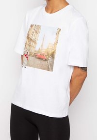 Vitt bomullströja med en grafisk tryck av en gata i Paris, med en röd bil och Eiffeltornet i bakgrunden. Kort ärm.