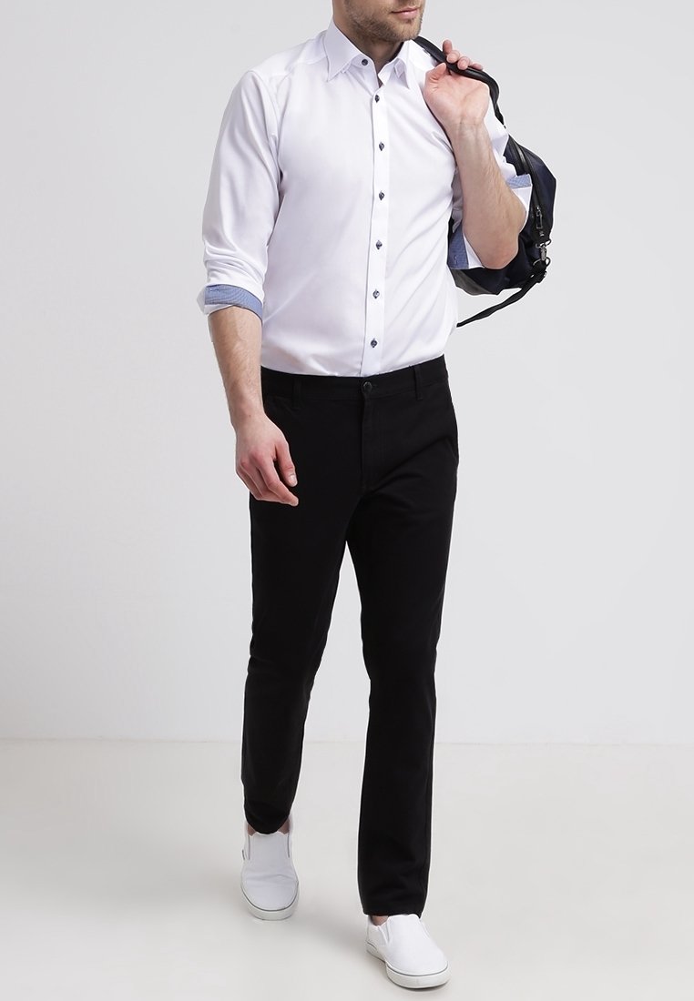 Homme marchant, portant une chemise blanche à boutons avec manches retroussées, pantalon noir, chaussures blanches sans lacets et portant un sac à dos noir sur une épaule.