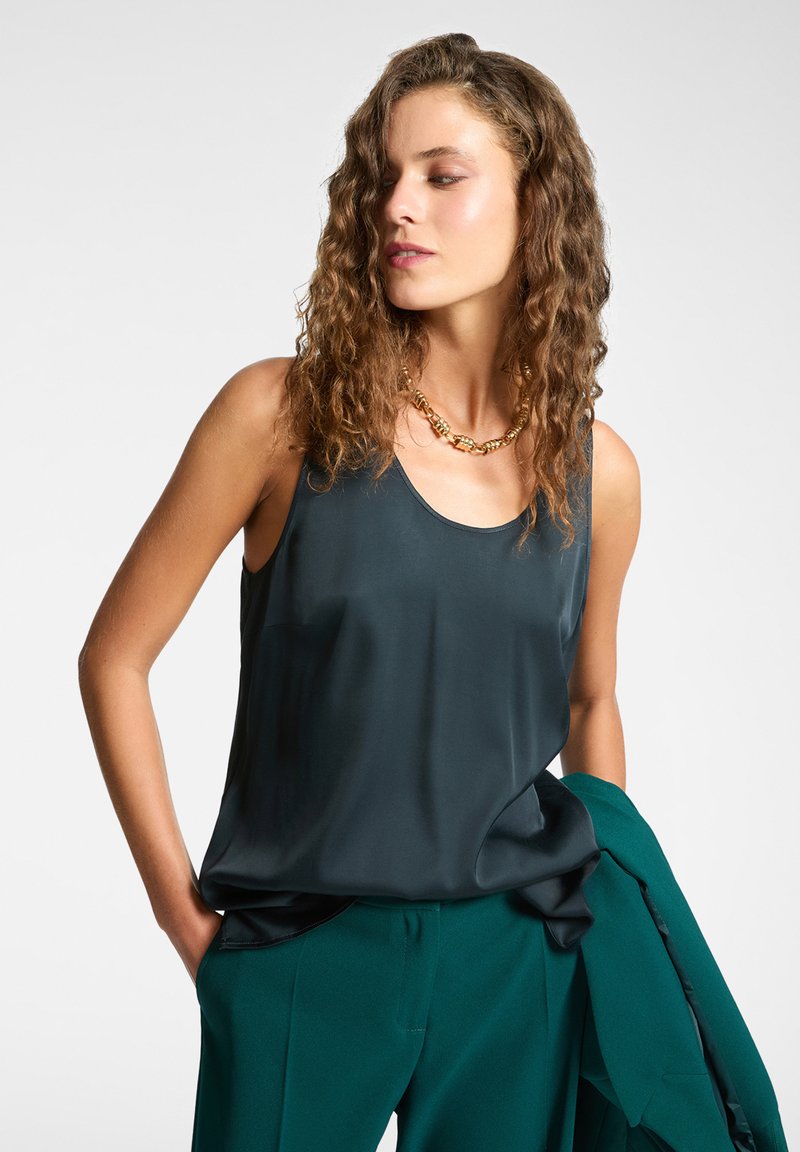 Top sin mangas de satén en verde azulado oscuro con un corte relajado, que presenta un escote redondo y una textura suave, combinado con pantalones verdes a medida.