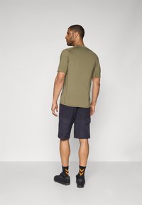 Chemise à manches courtes vert olive associée à un short cargo sombre. Chaussures noires avec des chaussettes à accent orange. Design simple et sportif avec une coupe décontractée.