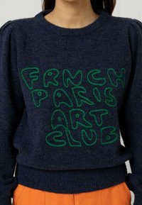 Pull bleu marine en matière douce avec un texte vert, texturé et surdimensionné : « FRNCH PARIS ART CLUB ». Manches bouffantes et taille cintrée.