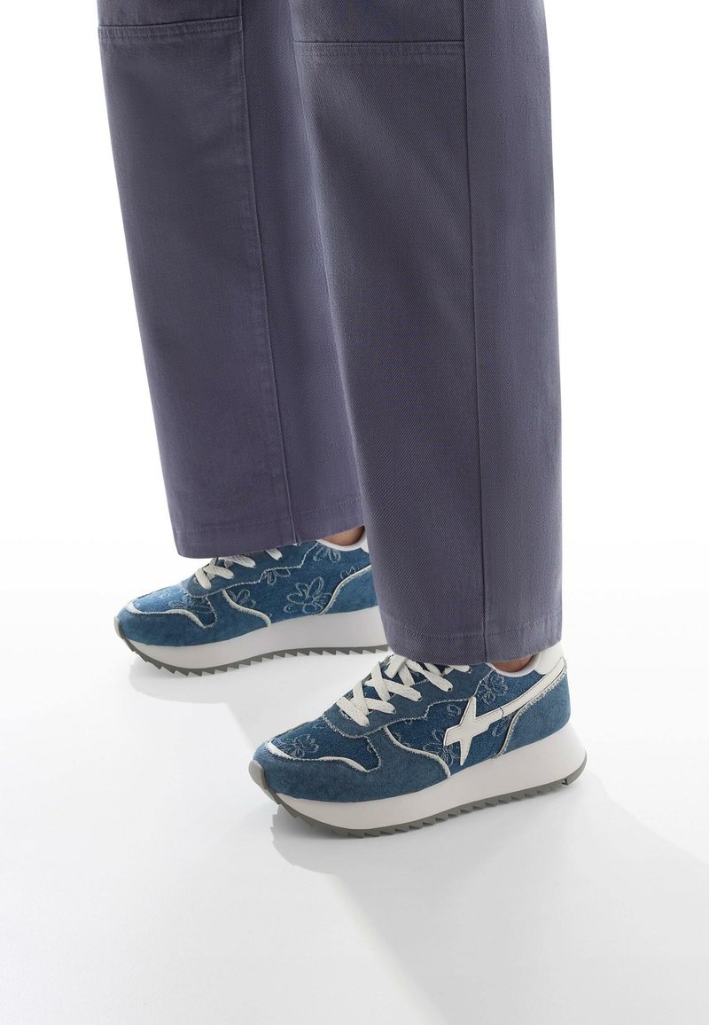 Sneaker sportive in denim blu con accenti bianchi, caratterizzati da ricami floreali, chiusura con lacci e una spessa suola bianca con fondo grigio testurizzato.