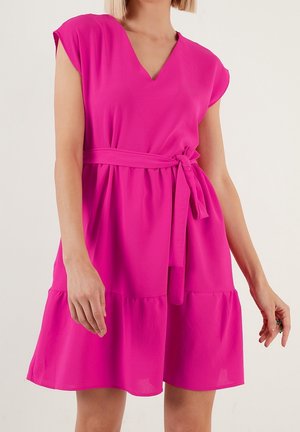 Femme portant une robe rose vif à col en V, manches courtes, ceinture nouée à la taille et ourlet à volants, debout devant un fond uni.