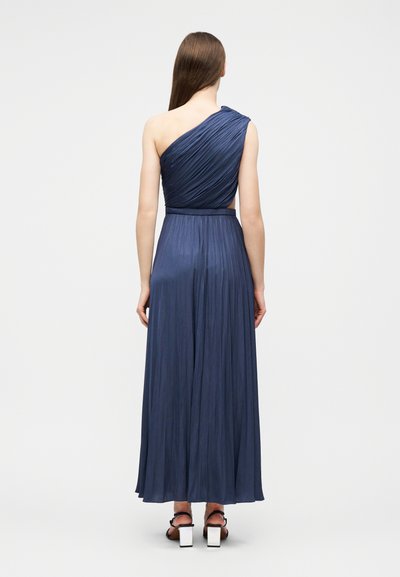 Femme aux longs cheveux bruns, vue de dos, portant une robe plissée bleu marine longueur au sol, à une épaule, et des sandales noires à talons blancs.