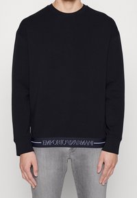 Svart sweatshirt med rund halsringning, långa ärmar och ett strukturerat logoband i marinblått vid fållen. Tillverkad i mjukt material.