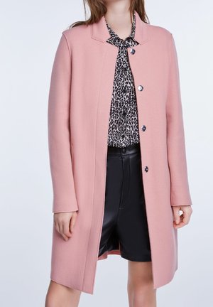 Donna che indossa un cappotto rosa fino al ginocchio sopra una camicetta con stampa leopardata in bianco e nero e pantaloncini neri a vita alta.