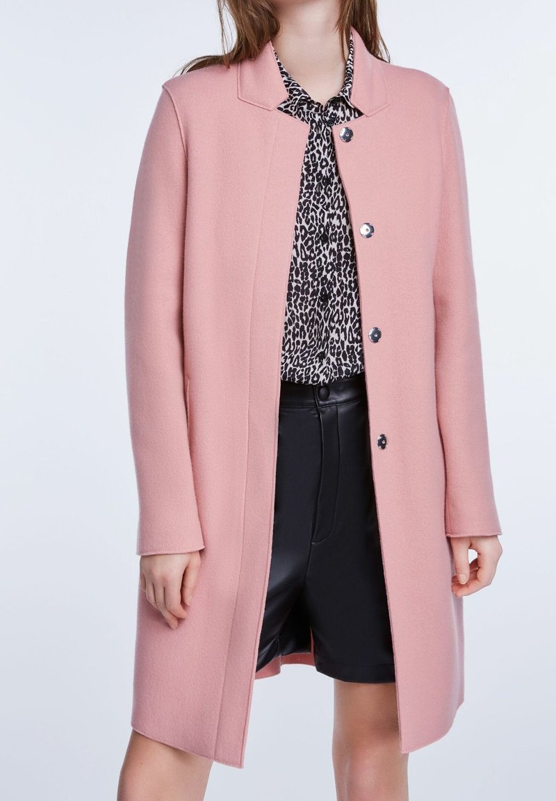 Femme portant un manteau rose jusqu'aux genoux sur un chemisier à imprimé léopard noir et blanc et un short taille haute noir.