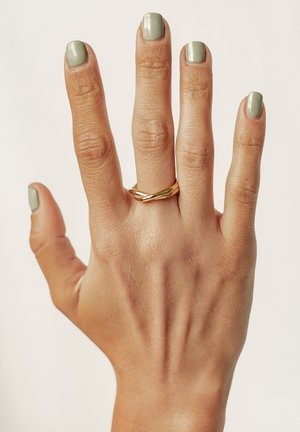 Anello d'oro intrecciato sull'anulare di una mano con pelle di tono chiaro e smalto verde su ciascun dito, su uno sfondo chiaro.