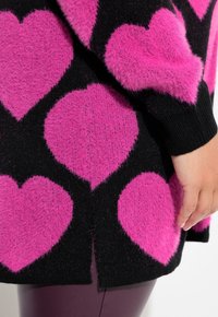 Schwarzer Pullover mit großem, wuscheligem rosa Herzmuster. Verfügt über gerippte Bündchen und Saum, körpernahe Länge mit seitlichen Schlitzdetails.