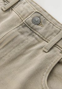 Nahaufnahme von beigem Denim-Stoff mit silbernem Knopf mit der Aufschrift "Premium Denim Goods" sowie sichtbaren Gürtelschlaufen und Nähten.