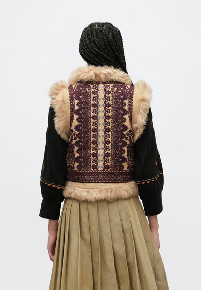 Γιλέκο από συνθετική γούνα με μωβ και μπεζ περίτεχνα σχέδια, με απαλό γιακά. Το άτομο φοράει πλισέ, ανοιχτό καφέ φούστα.