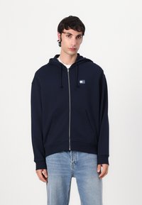 Marineblå zip-up hoodie med kængurulomme, justerbar snoretræk hætte og et lille logo patch på brystet. Fabrikater af bomuldsblanding.