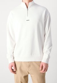 Maglione bianco con zip a quarto e colletto alto. Presenta polsini e orlo a coste, con un piccolo logo nero "HUGO" sul petto.