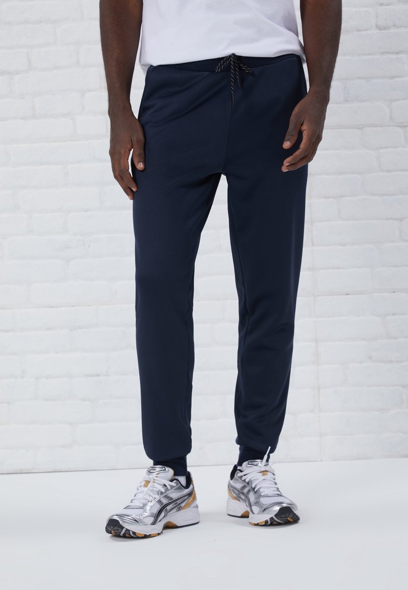 Námořnické modré sportovní kalhoty vyrobené z hladkého materiálu, s elastickým pasem a šňůrkou na stažení, doplněné stříbrnými a černými běžeckými botami.