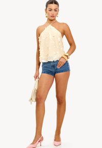 Femme portant un haut halter en sequins crème, un short en denim, des talons roses, tenant un sac à main crème, accessoirisée avec des boucles d'oreilles et des bracelets en or.