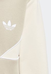 Ljusbeige sweatshirt med krämfärgade detaljer, med en vit trefoil-logotyp och pipade detaljer. Tillverkad av mjukt tyg med en texturerad yta.
