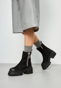 Stivaletti neri alla caviglia in suede con tacco spesso. Presentano una zip laterale con dettagli in argento e una suola testurizzata. Calze grigie visibili sopra gli stivaletti.