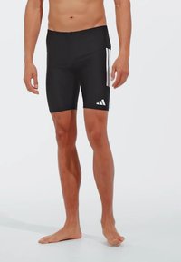 Crne uske kupaćice s glatkom teksturom, s tri bijele vertikalne pruge s jedne strane i Adidas logotipom blizu rubova.