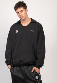 Schwarzes, übergroßes Sweatshirt mit Kragen, gesticktem Logo auf der Brust und strukturiertem Stoff. Kombiniert mit schwarzen Jogginghosen mit Kordelzug.