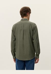 Groen geruite overhemd van stof met knoopmanchetten en een kraag. Heeft een rugplooi voor meer bewegingsvrijheid. Gemaakt met blauwe jeans.