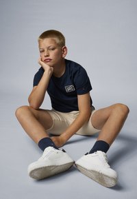 Marine T-skjorte med logo, beige shorts, marineblå sokker og hvite joggesko. Sittende stilling, kort blondt hår, lys hudtone. Nøytral bakgrunn.