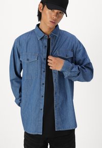 Camicia in denim con colletto, due tasche sul petto e chiusura a bottoni. Indossata sopra una maglietta nera, presenta una vestibilità rilassata e una tonalità blu.