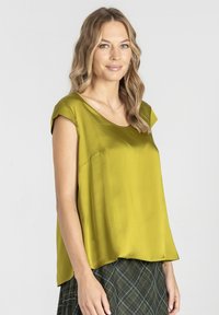 Kobieta z falującymi blond włosami, ubrana w błyszczącą oliwkową bluzkę na ramiączkach i ciemnozieloną spódnicę w kratę, stoi na jasnym tle.