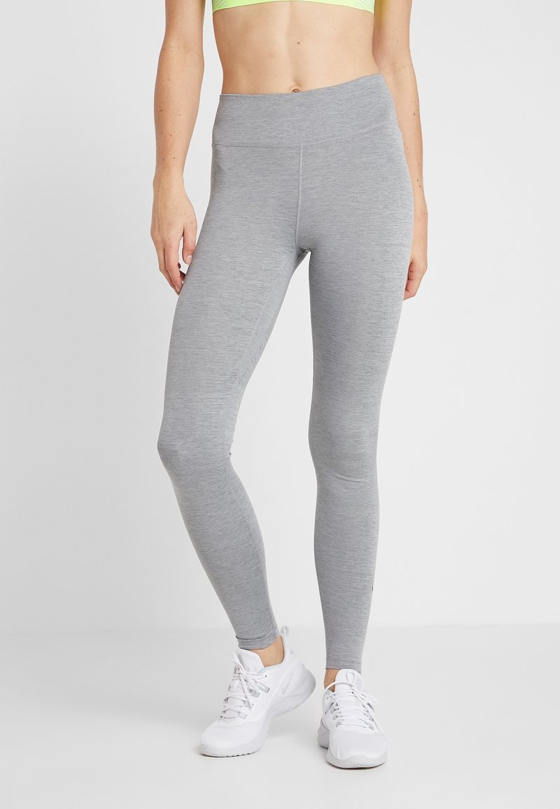 Femme portant des leggings ajustés gris clair, des chaussures de sport blanches et un haut court vert clair, debout sur un fond uni gris clair.