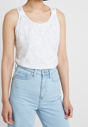 Woman wearing white sleeveless top with silver cactus prints tucked into light blue high-waisted jeans against a plain background.