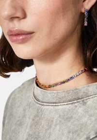 Collar en tono dorado con cuentas geométricas multicolores y pendientes a juego. Textura suave, acentos coloridos contra un suéter gris neutro.