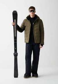 Slidžių įranga, kurioje yra juodos slidės su metaliniais akcentais ir tamsiai žalia pūkinė striukė, derinami su juodomis kelnėmis ir juodais slidinėjimo batais.