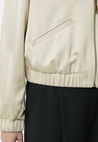 Beige Bomberjacke mit glatter Textur, elastischem Saum und Bündchen, ausgestattet mit einer seitlichen Tasche und einem metallischen Reißverschluss. Getragen über schwarzen Hosen.