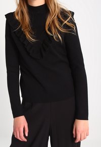 Svart långärmad topp med hög hals och volangdetalj fram, tillverkad av mjukt tyg med en slät textur.