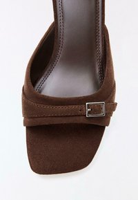 Sandale ouverte en daim marron avec bout carré, bride traversant le pied maintenue par une petite boucle argentée, et semelle intérieure en cuir lisse.