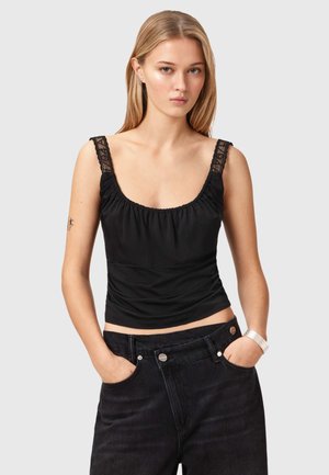 Vrouw met lang blond haar, een zwart mouwloos topje met kanten bandjes en zwarte high-waisted jeans, handen in de zakken.