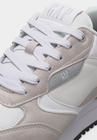 Gros plan sur une chaussure de sport blanche et beige avec des lacets blancs et le logo GAP sur la languette et le panneau latéral.