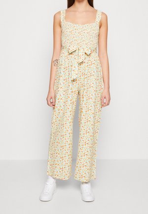 Frau in einem ärmellosen, karierten Jumpsuit mit gerafftem Oberteil, weitem Bein und einer Schleife an der Taille, kombiniert mit weißen Sneakers.