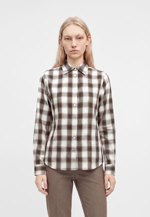 Femme aux longs cheveux blonds portant une chemise à carreaux marron et blanc boutonnée et un pantalon marron, debout devant un fond clair uni.