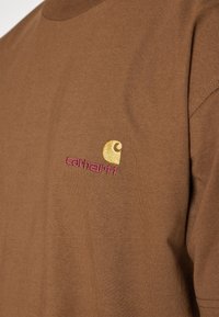 Camiseta de algodón marrón con un pequeño logotipo bordado de Carhartt en amarillo y rosa en el lado izquierdo del pecho. Diseño simple y casual.