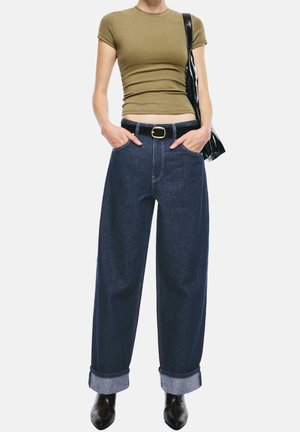 Osoba ubrana w oliwkowo zielony dopasowany T-shirt, ciemnoniebieskie dżinsy o szerokich nogawkach z mankietami i czarnym paskiem, czarne botki oraz czarną torbę na ramię, ręce w kieszeniach.