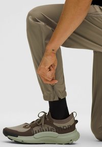 Beige sportbroeken met elastische manchetten en ritsen, gecombineerd met bruine en groene sneakers met een bovenkant van mesh en geprinte accenten.
