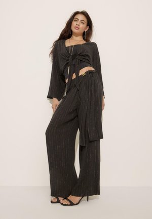 Žena oblečená v čiernych širokých nohaviciach a zodpovedajúcom zaväzovanom crop tope s tenkými zlatými vertikálnymi pruhmi, stojaca na svetlom jednolennom pozadí.