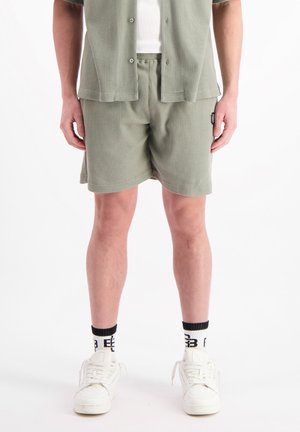 Olijfgroene, gestructureerde short-sleeve shirt en bijpassende shorts in geribbeld materiaal. Witte tanktop eronder, gecombineerd met witte sneakers en zwarte sokken.